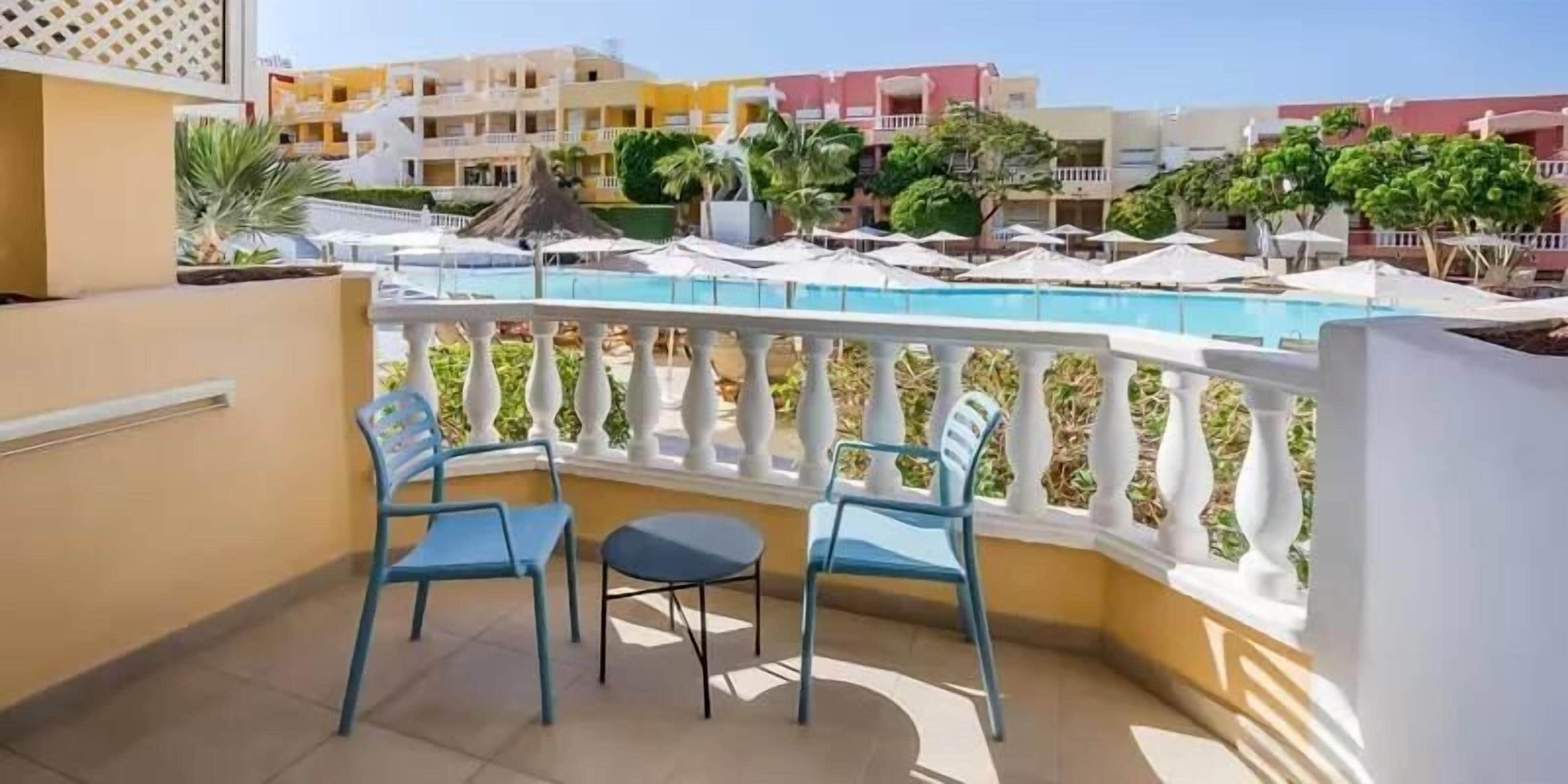 A balcony with two chairs and a small table, overlooking a pool area with umbrellas and lounge chairs.