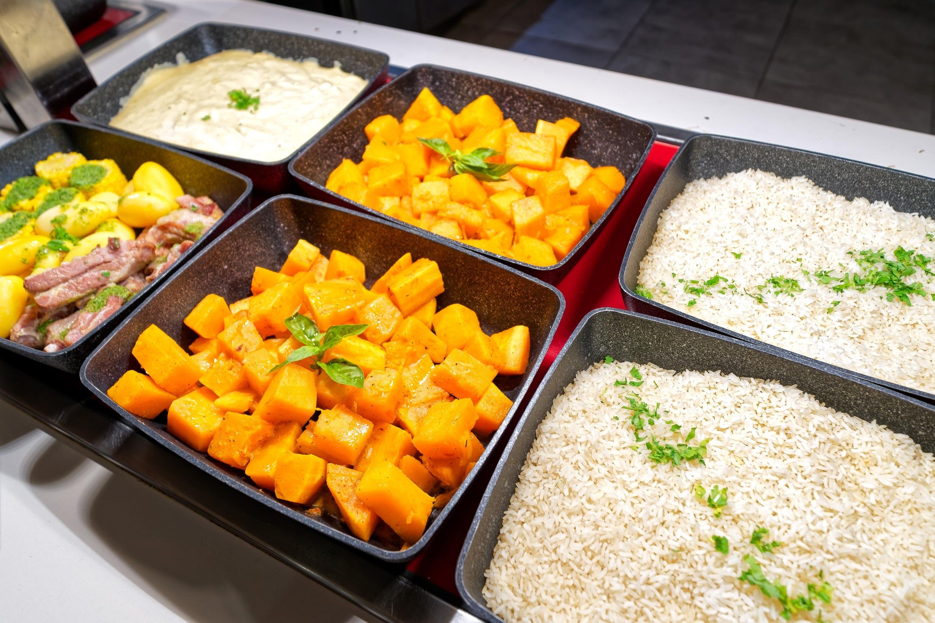 Buffet com diversas opções de comida, incluindo arroz, vegetais em cubos e um prato cremoso.