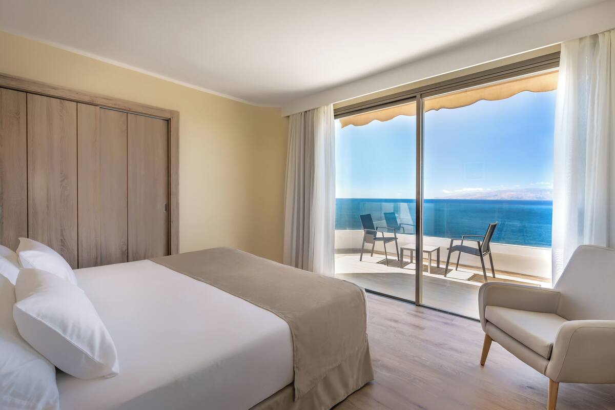 A hotel room with a bed, a chair, and a balcony with a view of the sea.