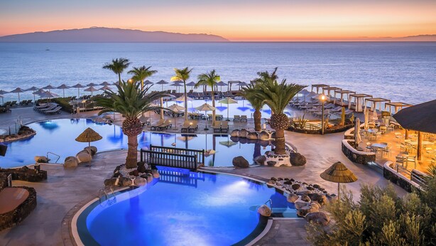 A resort pool area with lounge chairs, umbrellas, and a view of the sea at sunset. - Barceló Santiago - Adults Only (Santiago del Teide)