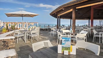 Un patio al aire libre con mesas y sillas, vista al mar y una zona de estar cubierta.