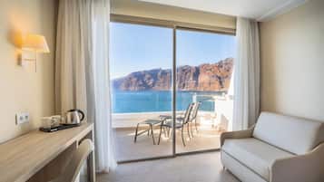 A hotel room with a balcony, a sofa, a desk with a kettle, and a view of the sea and cliffs.