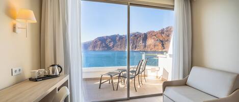 A hotel room with a balcony, a sofa, a desk with a kettle, and a view of the sea and cliffs.