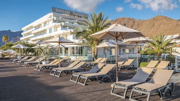 Un complejo turístico con numerosas sillas de descanso y sombrillas en una terraza de madera, un edificio con un cartel y palmeras.