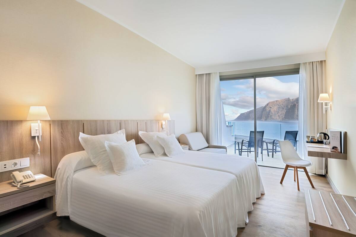 A hotel room with two beds, a desk, a chair, and a view of the sea.