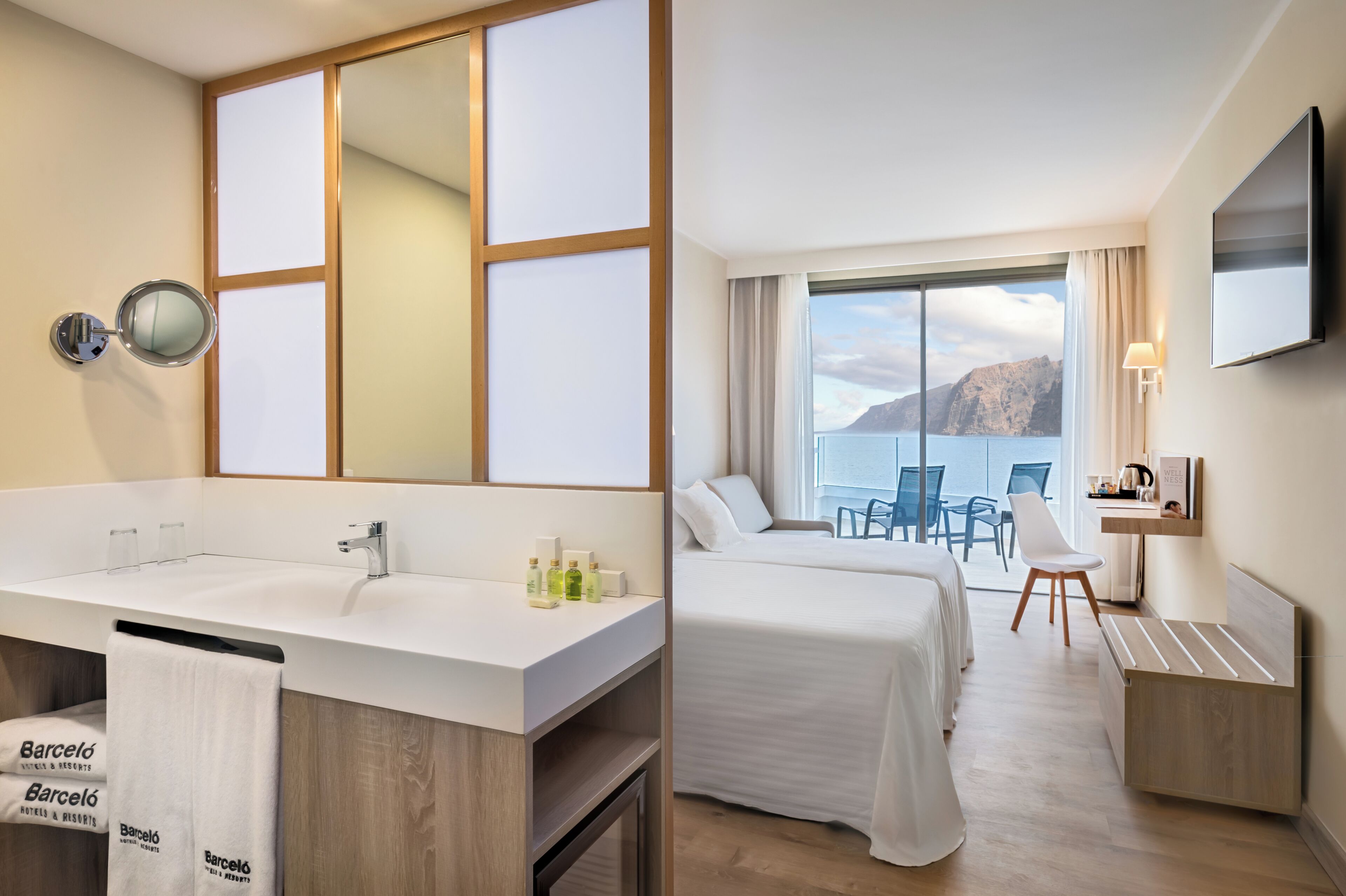 A hotel room with a large mirror, a sink, a bed, a TV, and a balcony with a view.