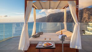 A balcony with a canopy, a bed, a table with a plate of fruit and two glasses of wine, and a view of the sea and cliffs. - Barceló Santiago - Adults Only (Santiago del Teide)