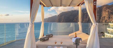 A balcony with a canopy, a bed, a table with a plate of fruit and two glasses of wine, and a view of the sea and cliffs.