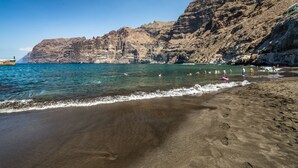 A beach with clear blue water, rocky cliffs, and footprints in the sand. - Barceló Santiago - Adults Only (Santiago del Teide)