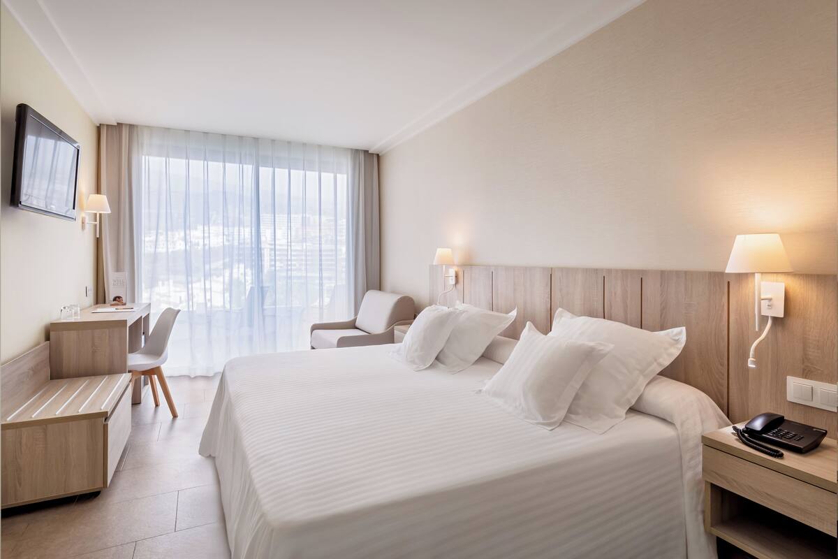 A hotel room with a large bed, a desk with a chair, a TV mounted on the wall, and a window with curtains.