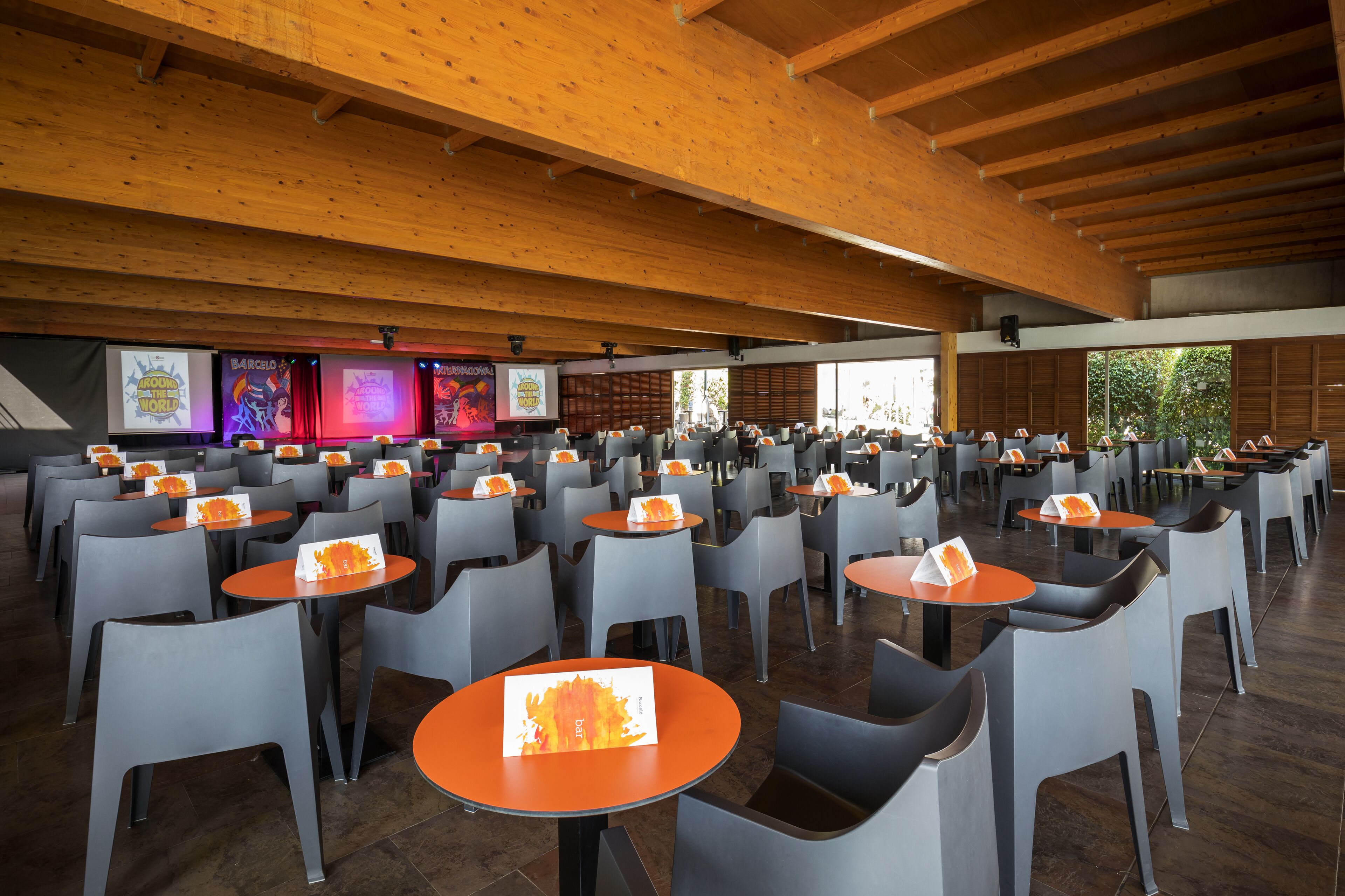 A spacious indoor area with multiple tables and chairs, wooden beams on the ceiling, and large windows allowing natural light.