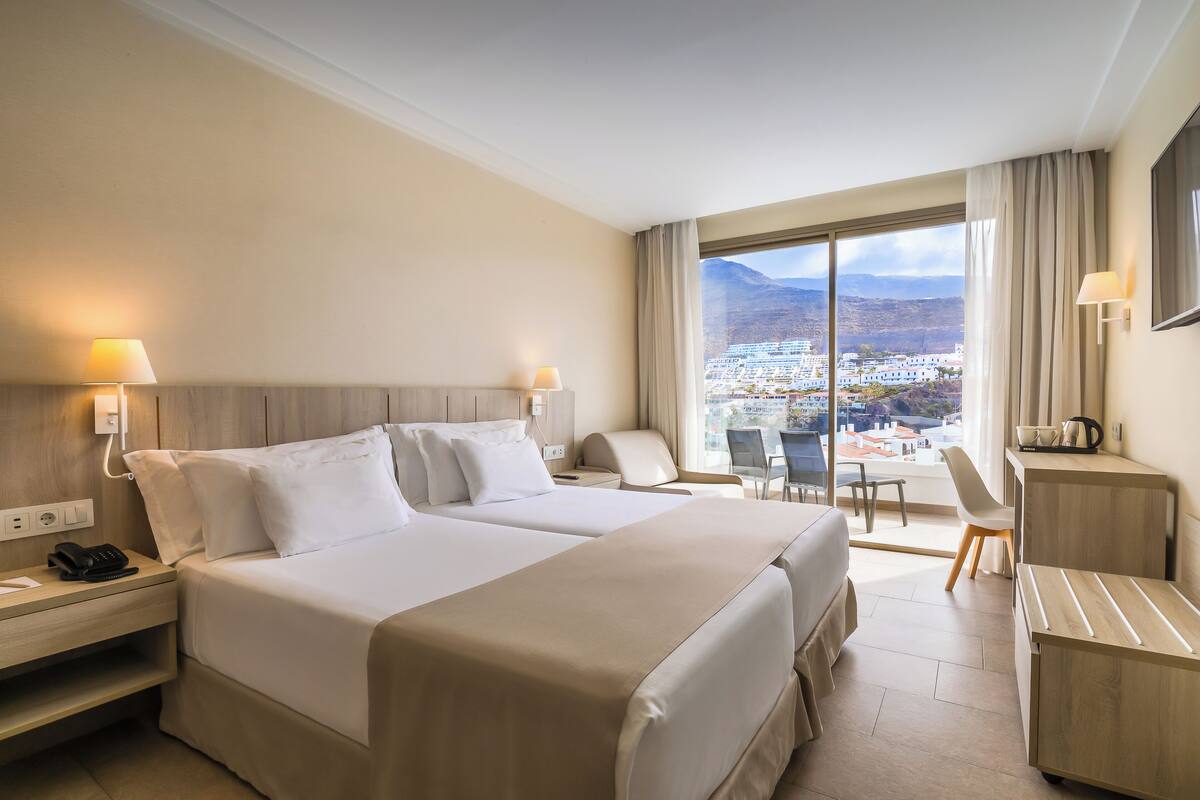 A hotel room with two beds, a desk, a chair, and a view of a cityscape.