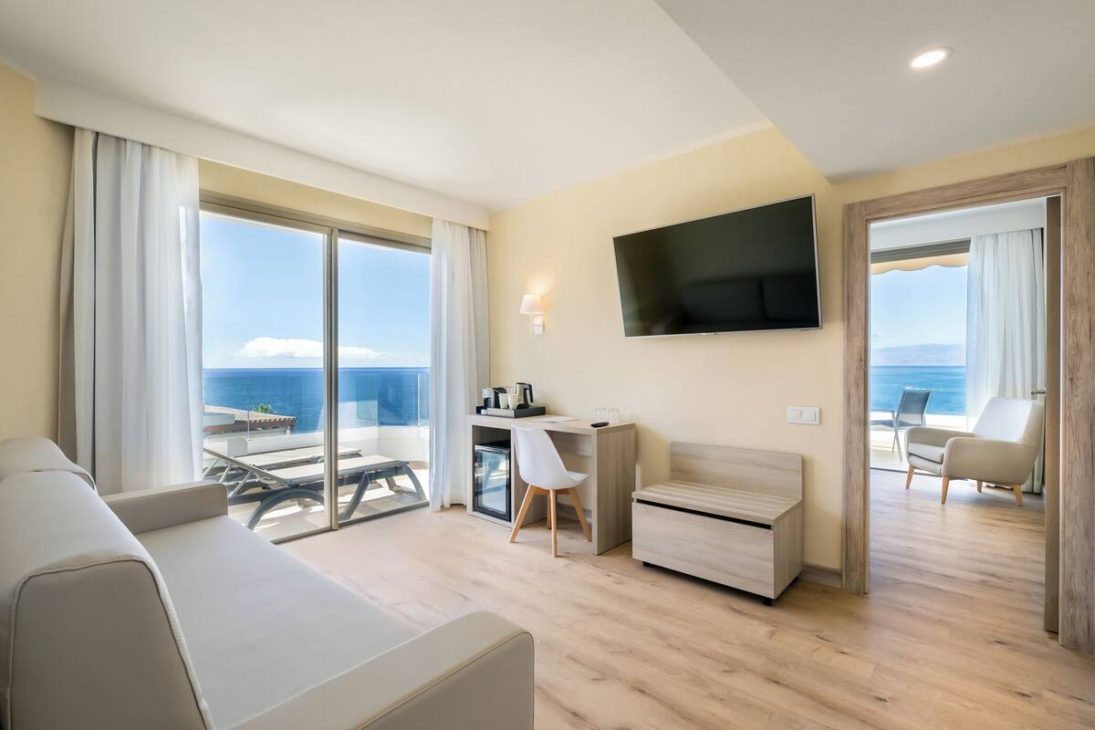 A modern hotel room with a large balcony, a flat-screen TV, a desk, a sofa, and a chair.