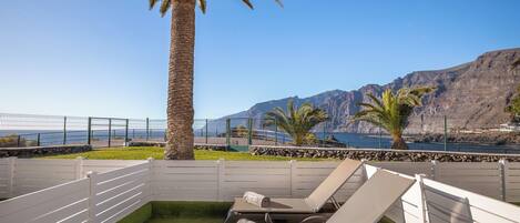 A patio with two lounge chairs, a palm tree, and a view of the sea and mountains.
