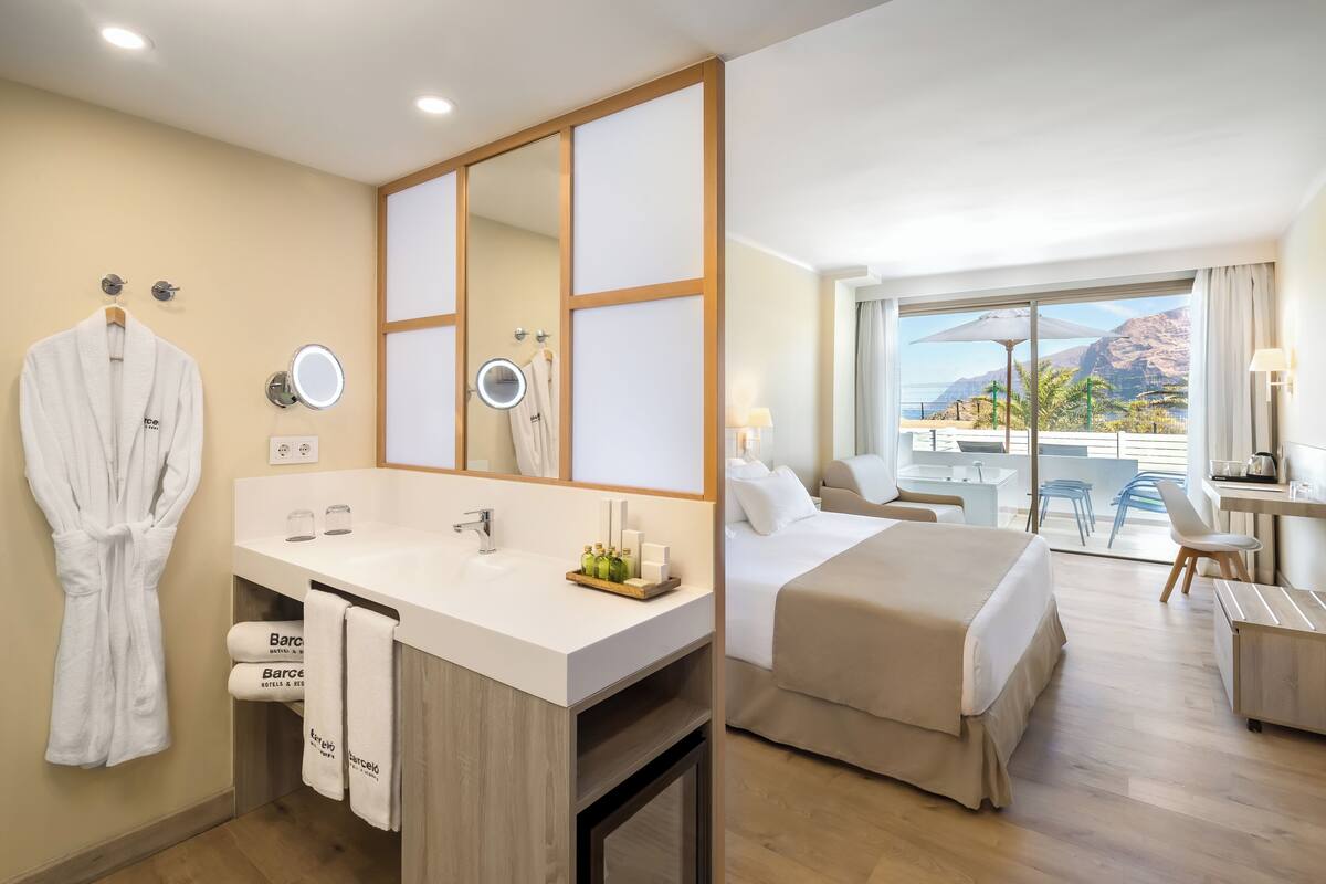 A hotel room with a large mirror, a sink with toiletries, a bed, a desk, and a view of the outdoors.