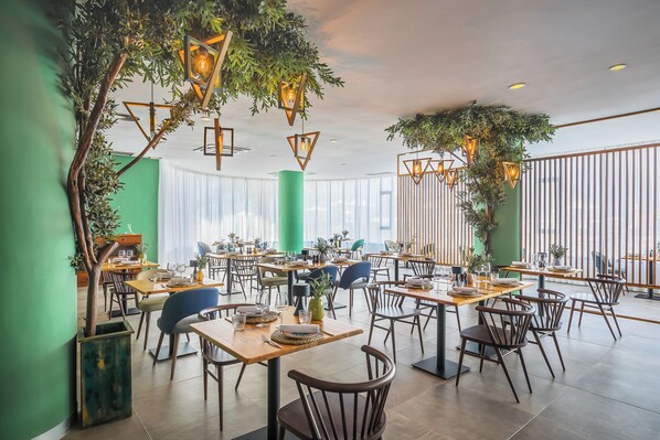 Un comedor amplio con varias mesas y sillas, ventanas grandes y plantas colgantes.