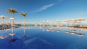 Una piscina infinita azul cristalina con sillones de descanso y sombrillas, que refleja el cielo y las palmeras.