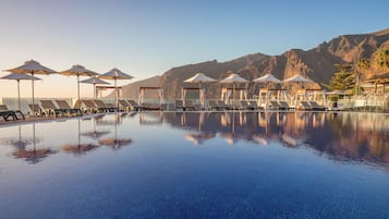 Una piscina con sillones reclinables y sombrillas, rodeada de montañas.