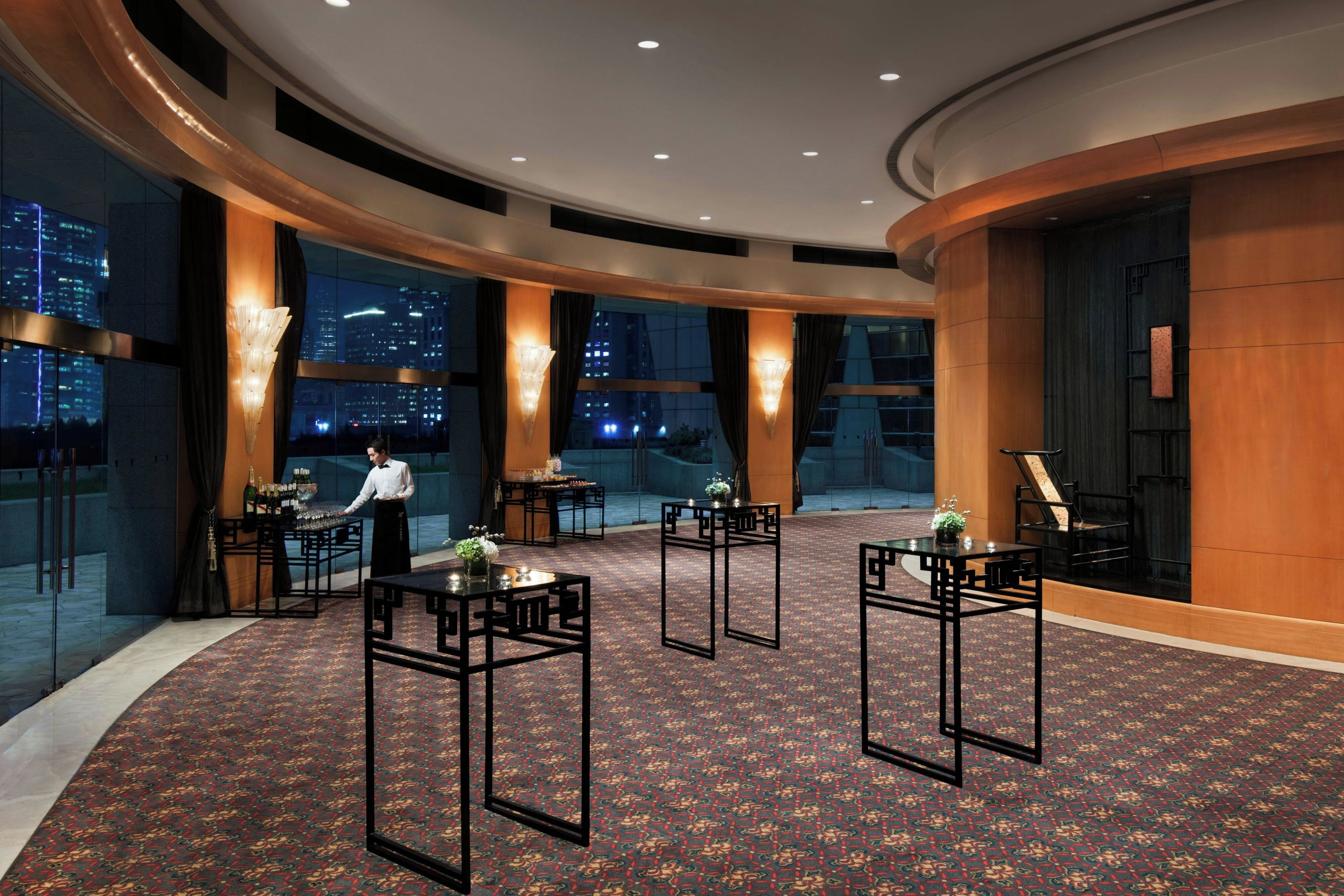 Un vestíbulo de hotel con distribución circular, grandes ventanales que ofrecen vistas a la ciudad y varias mesas con sillas.