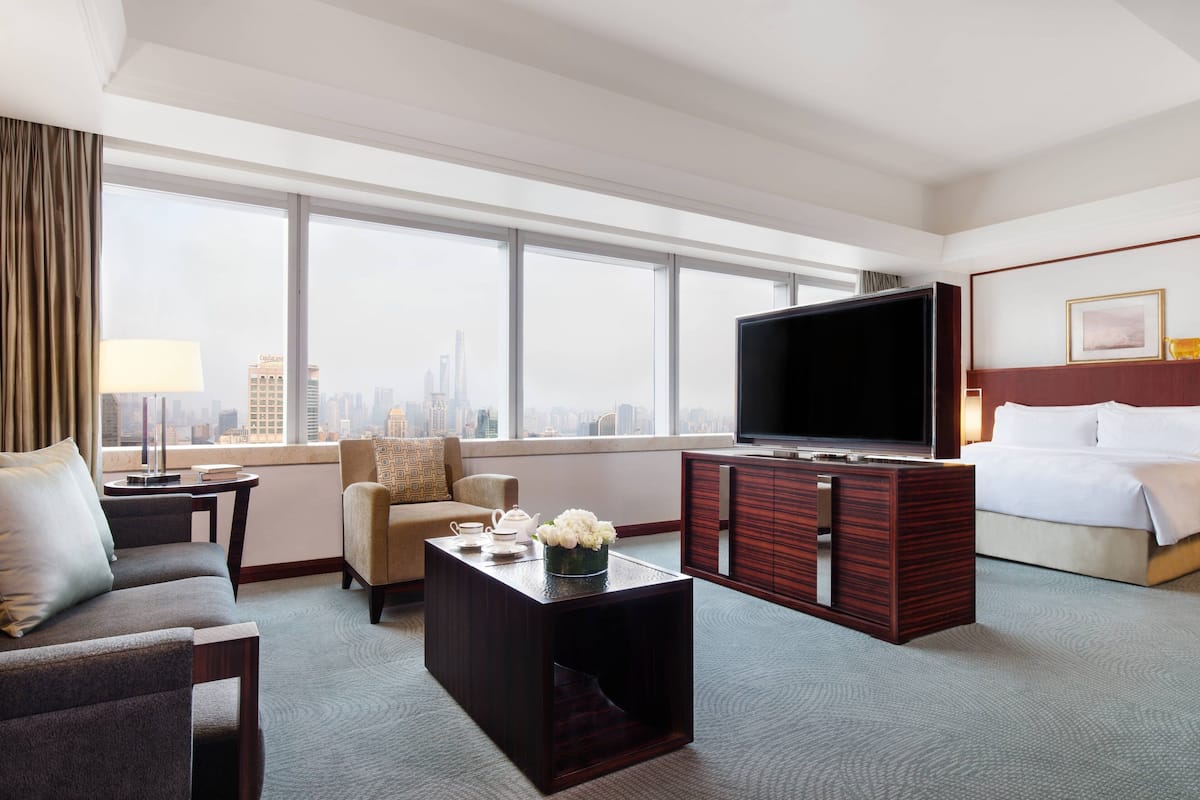 A hotel room with a large window, a flat-screen TV, a sofa, a coffee table, a bed, and a dresser.