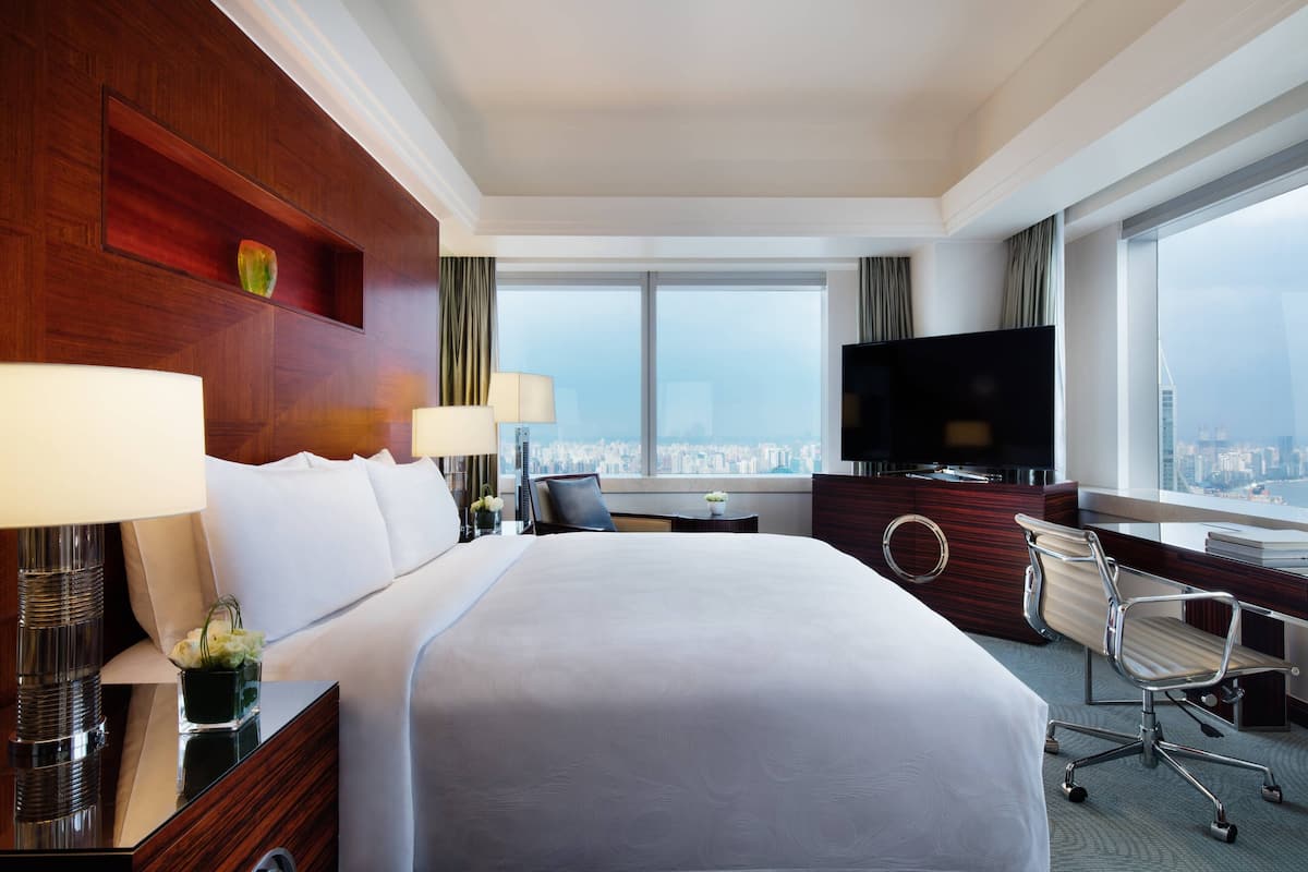 A hotel room with a large bed, a desk, a chair, a TV, and a view of the city.