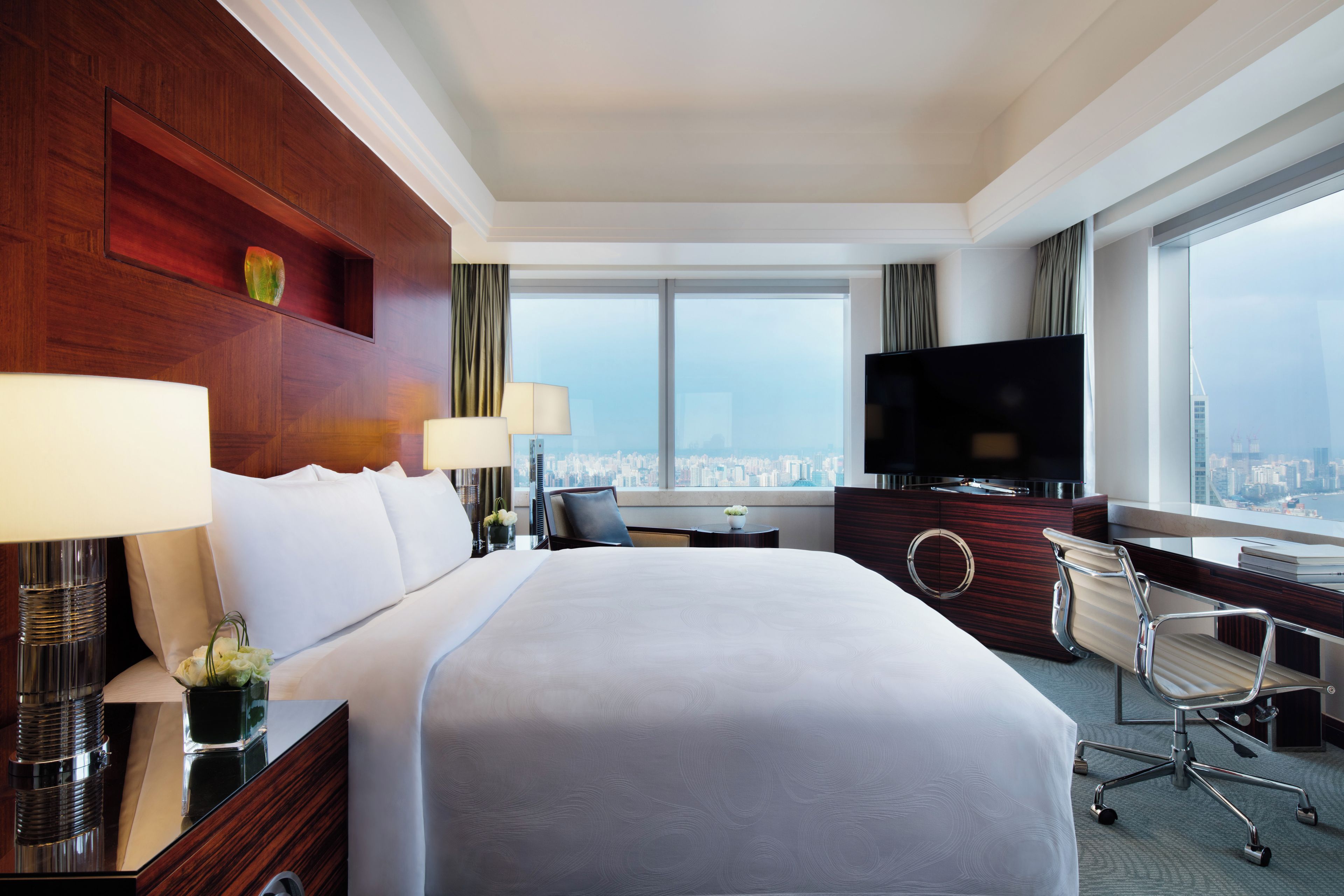 A hotel room with a large bed, a desk, a chair, a television, and a view of a cityscape through large windows.