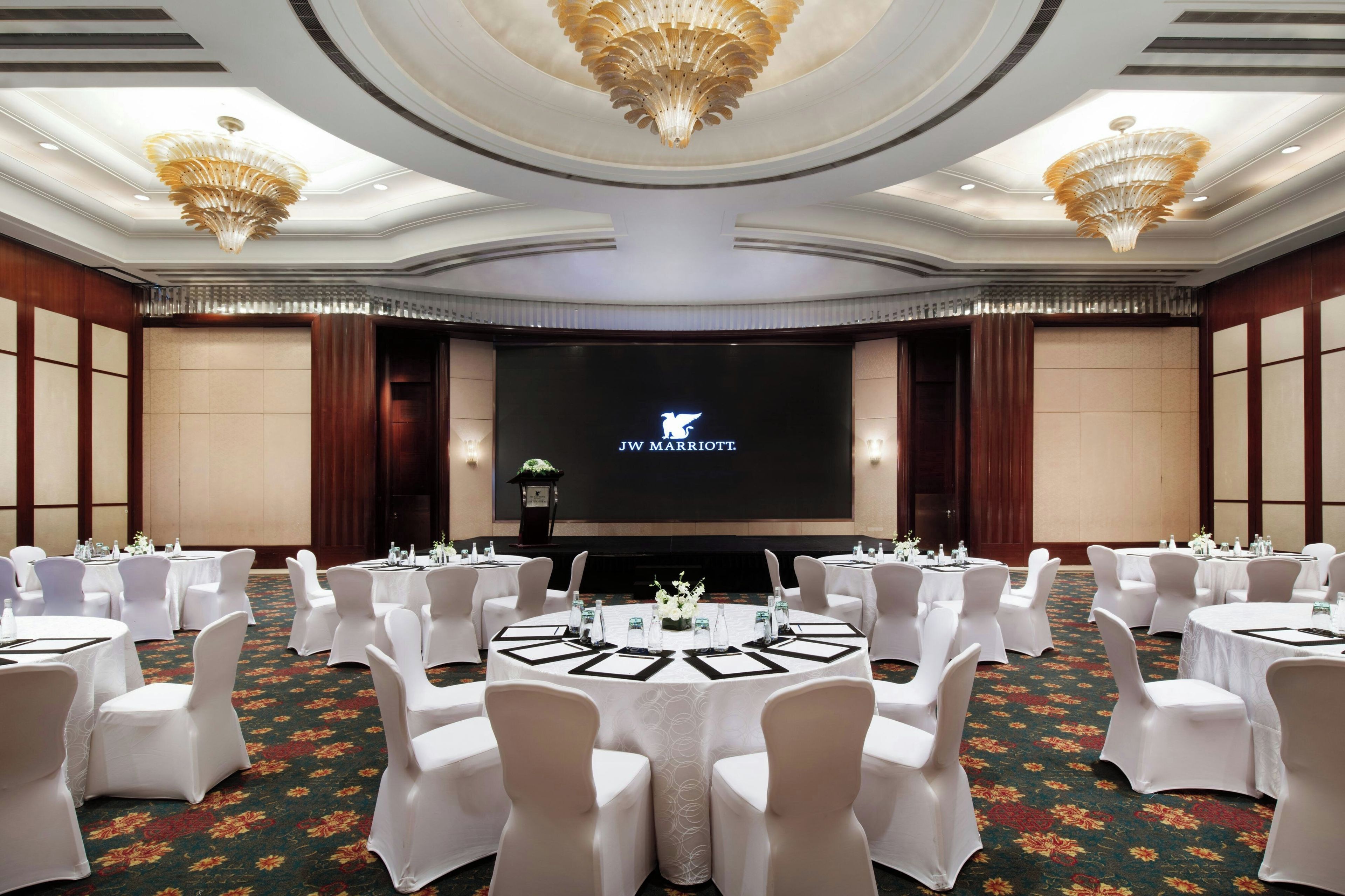 A JW Marriott conference room with round tables, white chairs, and a large screen displaying the hotel logo.