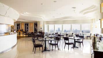 A spacious restaurant with high ceilings and large windows offering city views. Tables are neatly arranged with chairs.