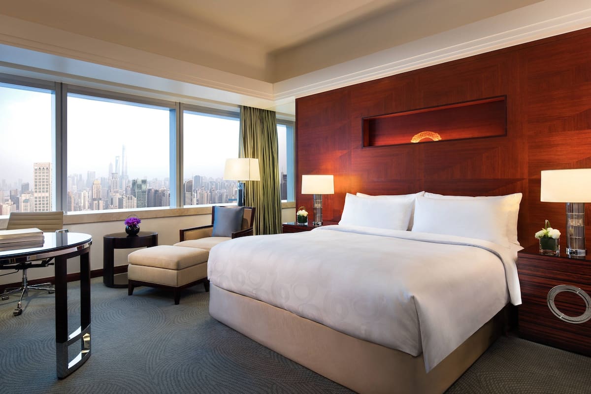 A hotel room with a large bed, two bedside tables with lamps, a desk with a chair, a view of the city through large windows, and a cityscape visible outside.