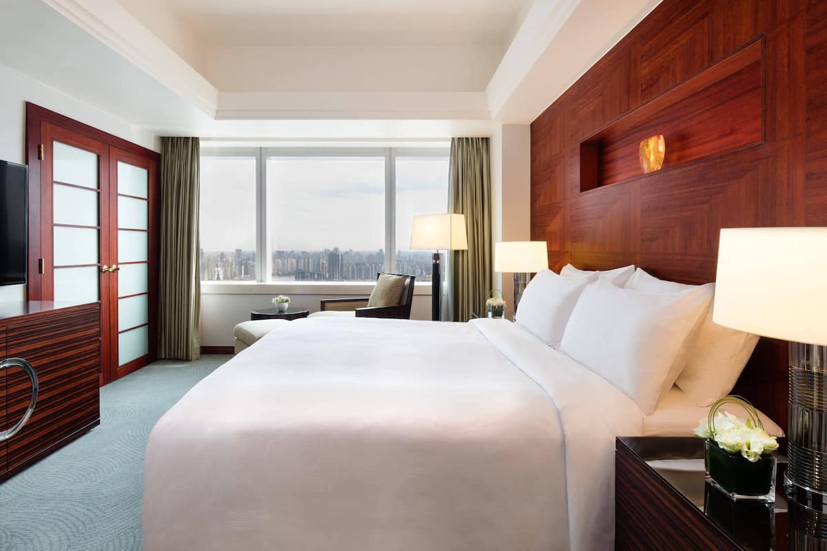 A hotel room with a large bed, a television, a desk, and a window with a city view.