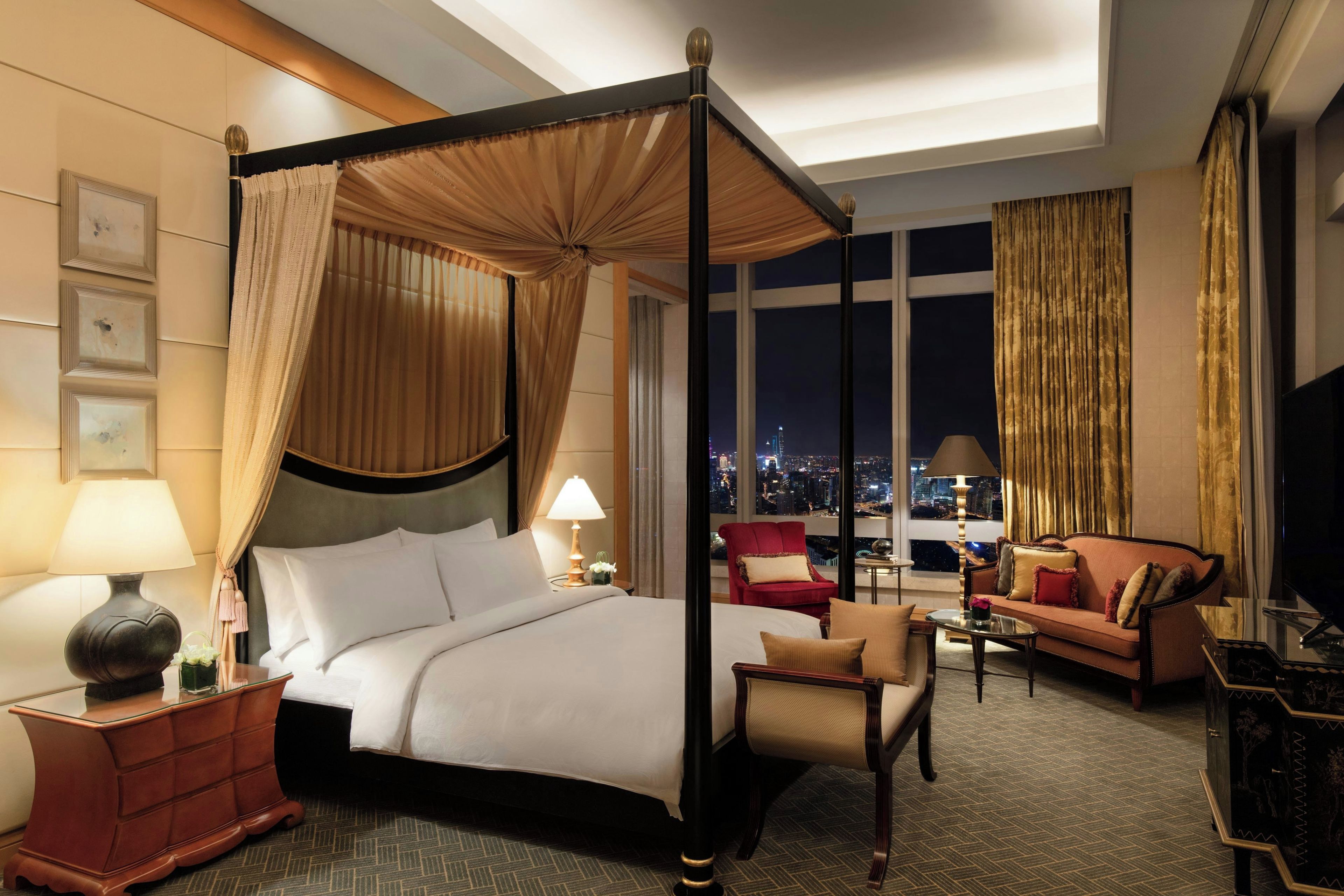 A four-poster bed with a canopy, a red armchair, a sofa, and a television in a hotel room with a city view.