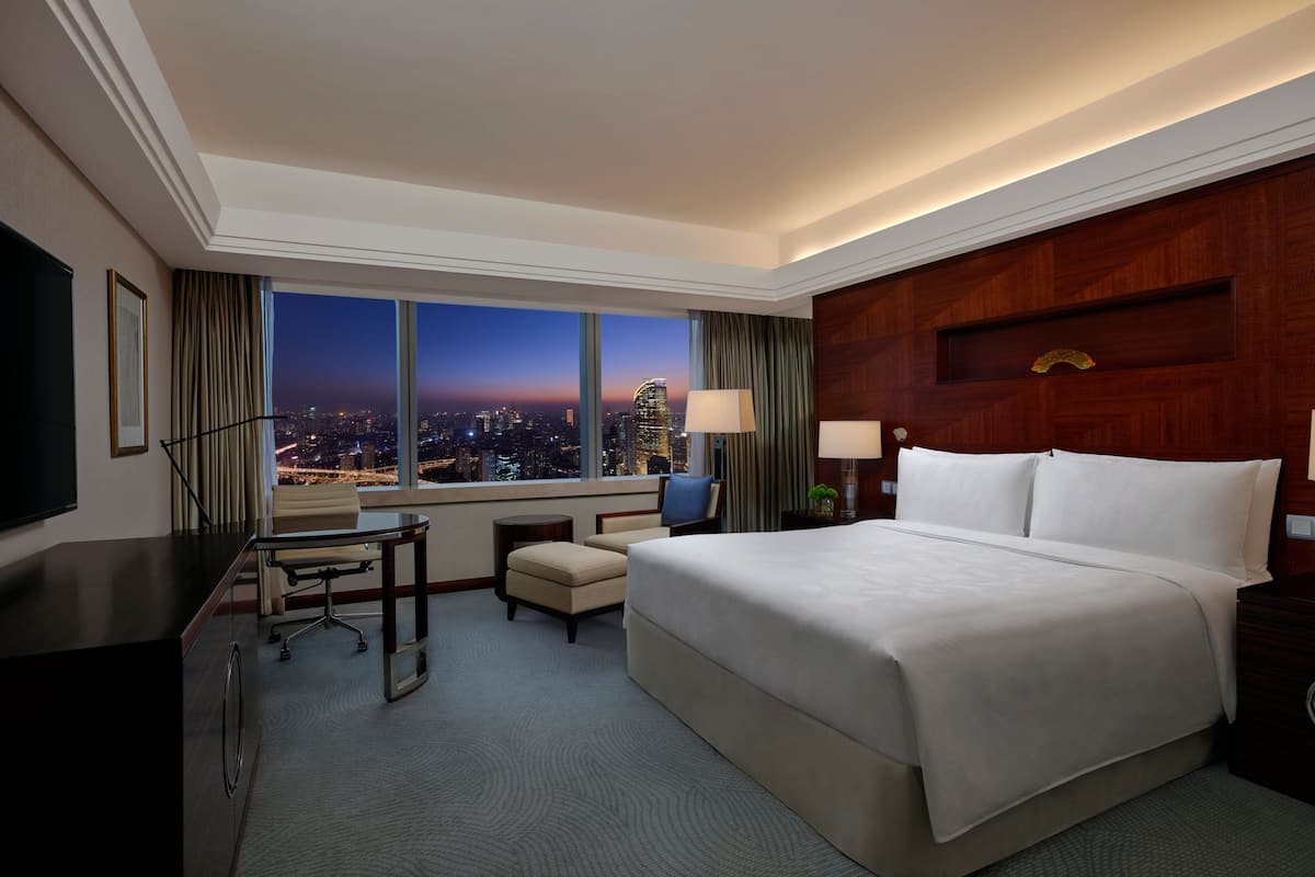 A hotel room with a large bed, a desk, a chair, a TV, and a view of the city at dusk.
