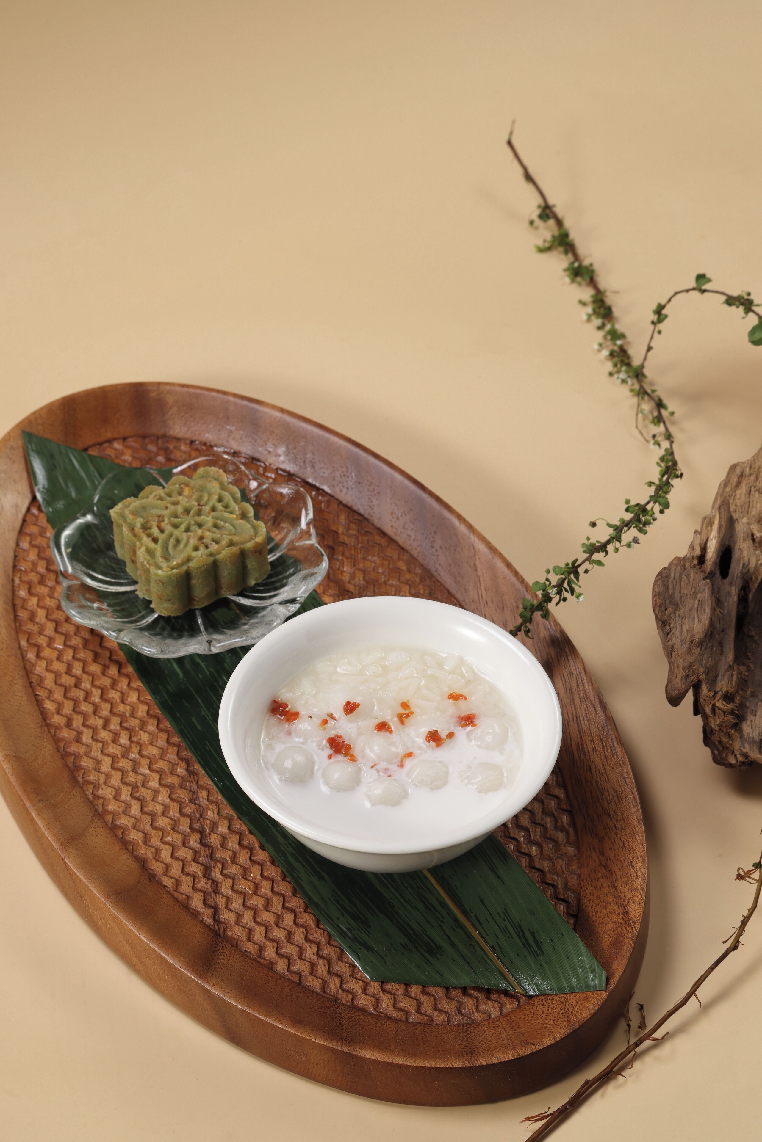 A wooden tray with a bowl of white rice topped with red chili flakes and a decorative green cake on a woven mat.