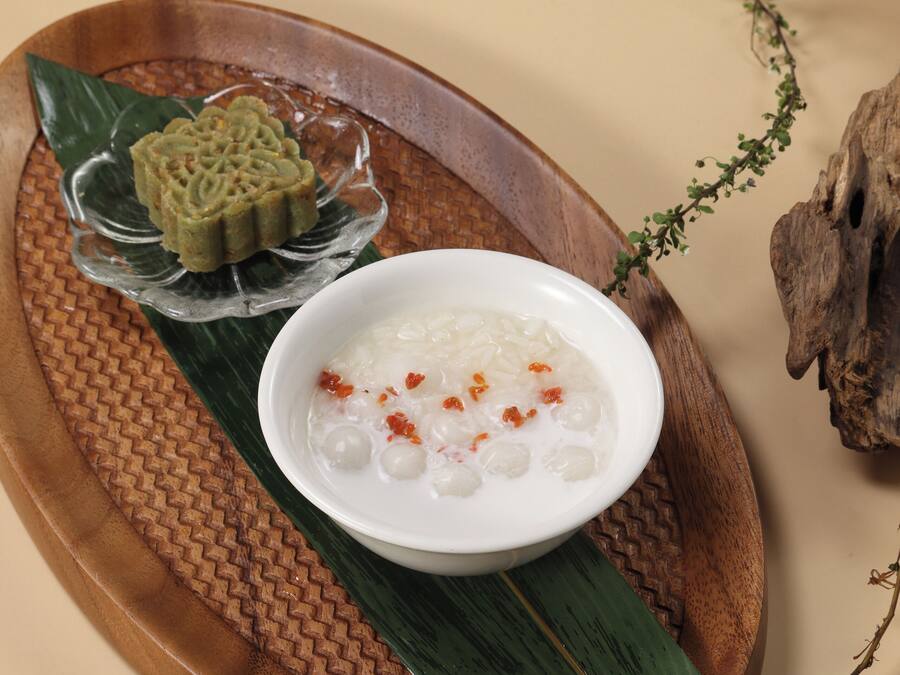 A wooden tray with a bowl of white rice topped with red chili flakes and a decorative green cake on a woven mat.