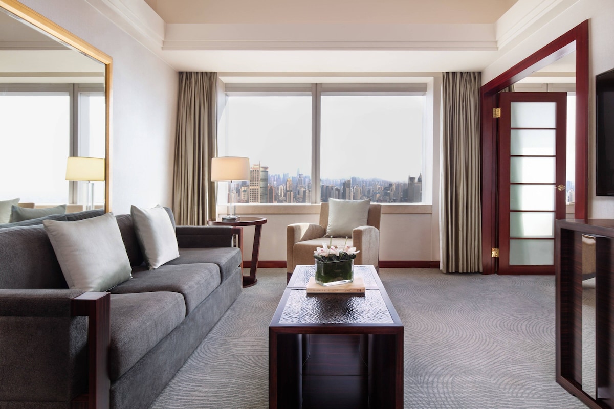 A hotel room with a grey sofa, a small table with a vase, a chair, and a large window with a city view.