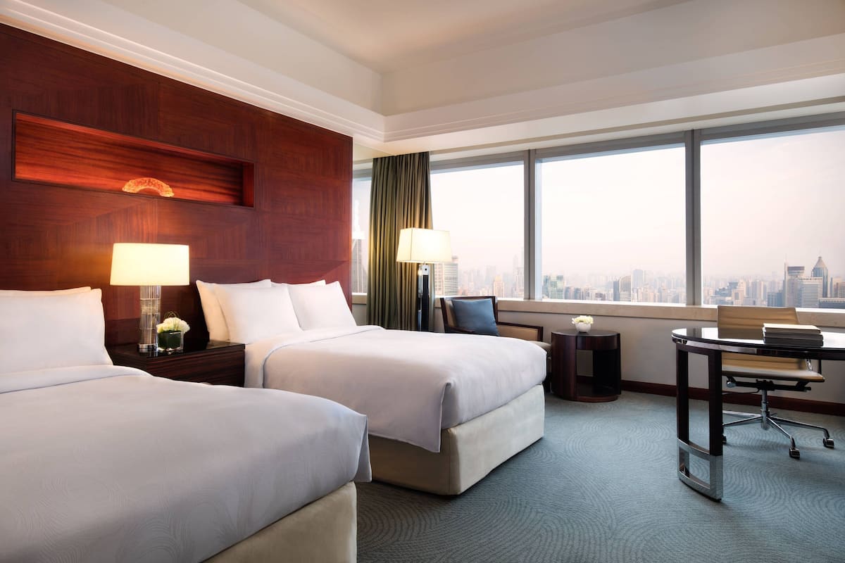 A hotel room with two beds, a desk, a chair, and a view of the city.