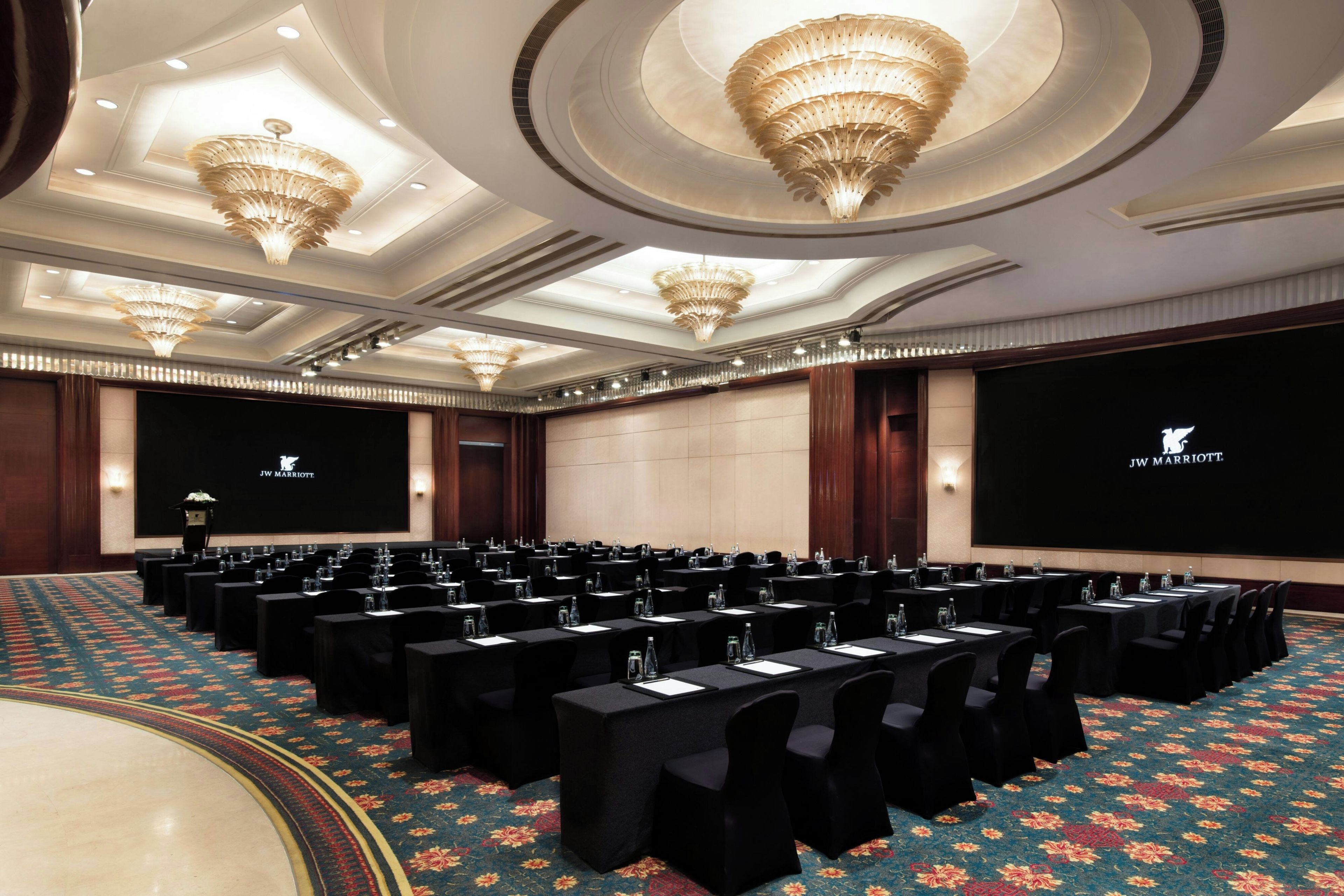 A conference room with rows of tables, black chairs, and a large screen displaying the JW Marriott logo.