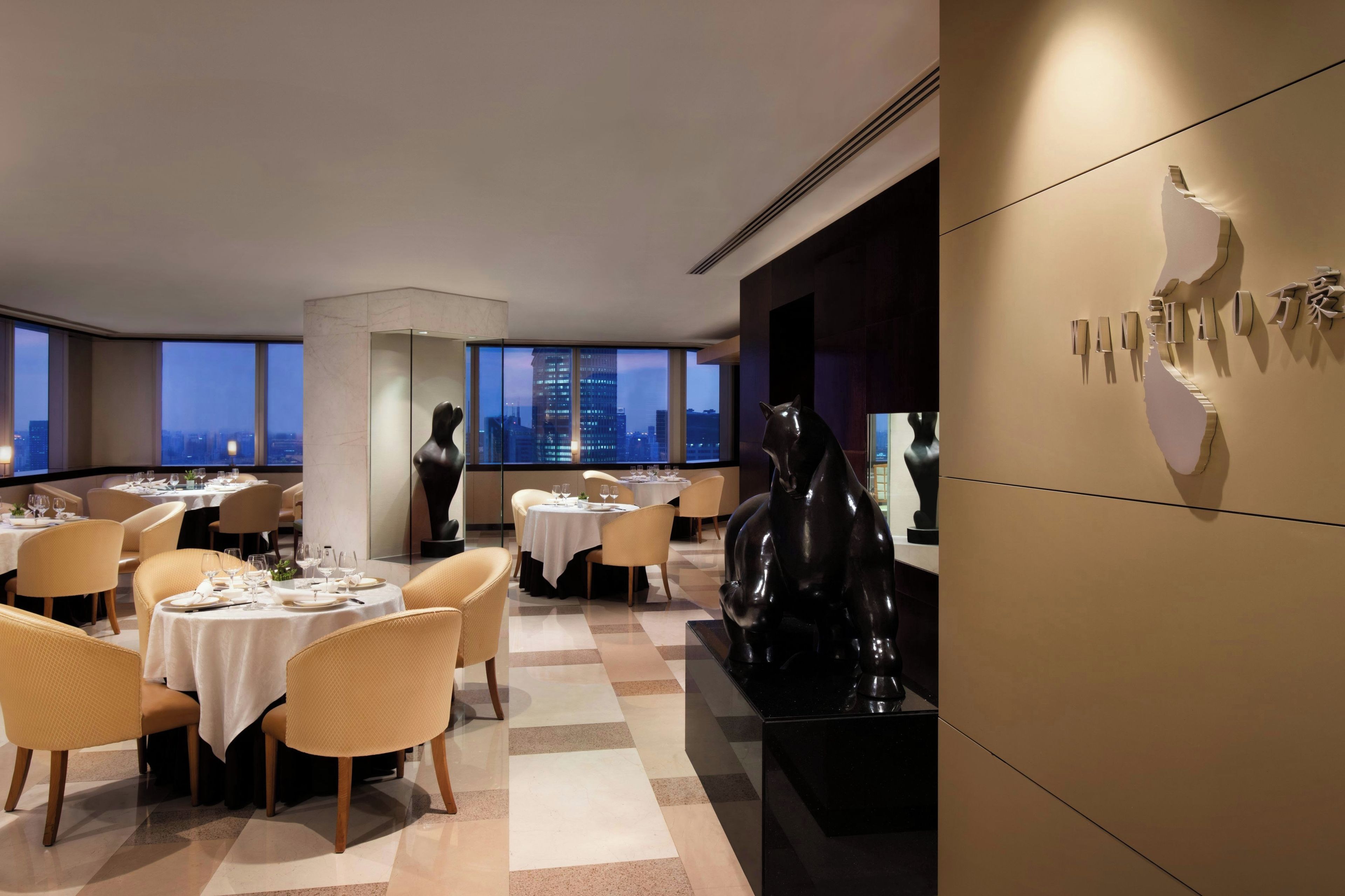 Un comedor moderno con mesas dispuestas para comensales, amplias ventanas que ofrecen vistas a la ciudad y una escultura negra distintiva en la pared.