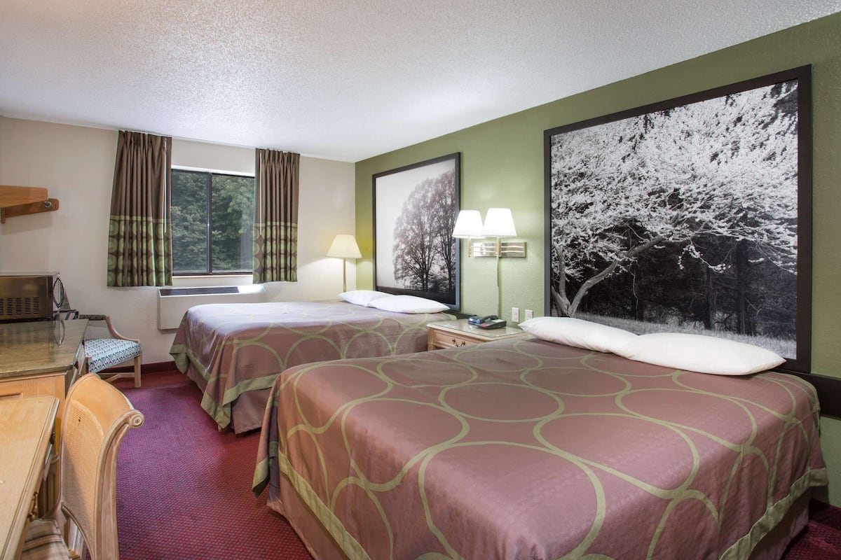 Une chambre d’hôtel avec deux lits, un bureau, une chaise, une fenêtre avec des rideaux et un grand tableau encadré au mur.