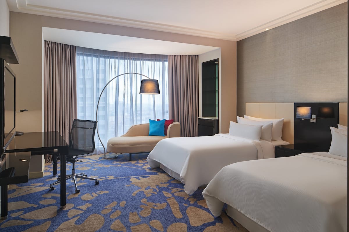 A hotel room with two beds, a desk, a chair, a large window with curtains, and a floor lamp.