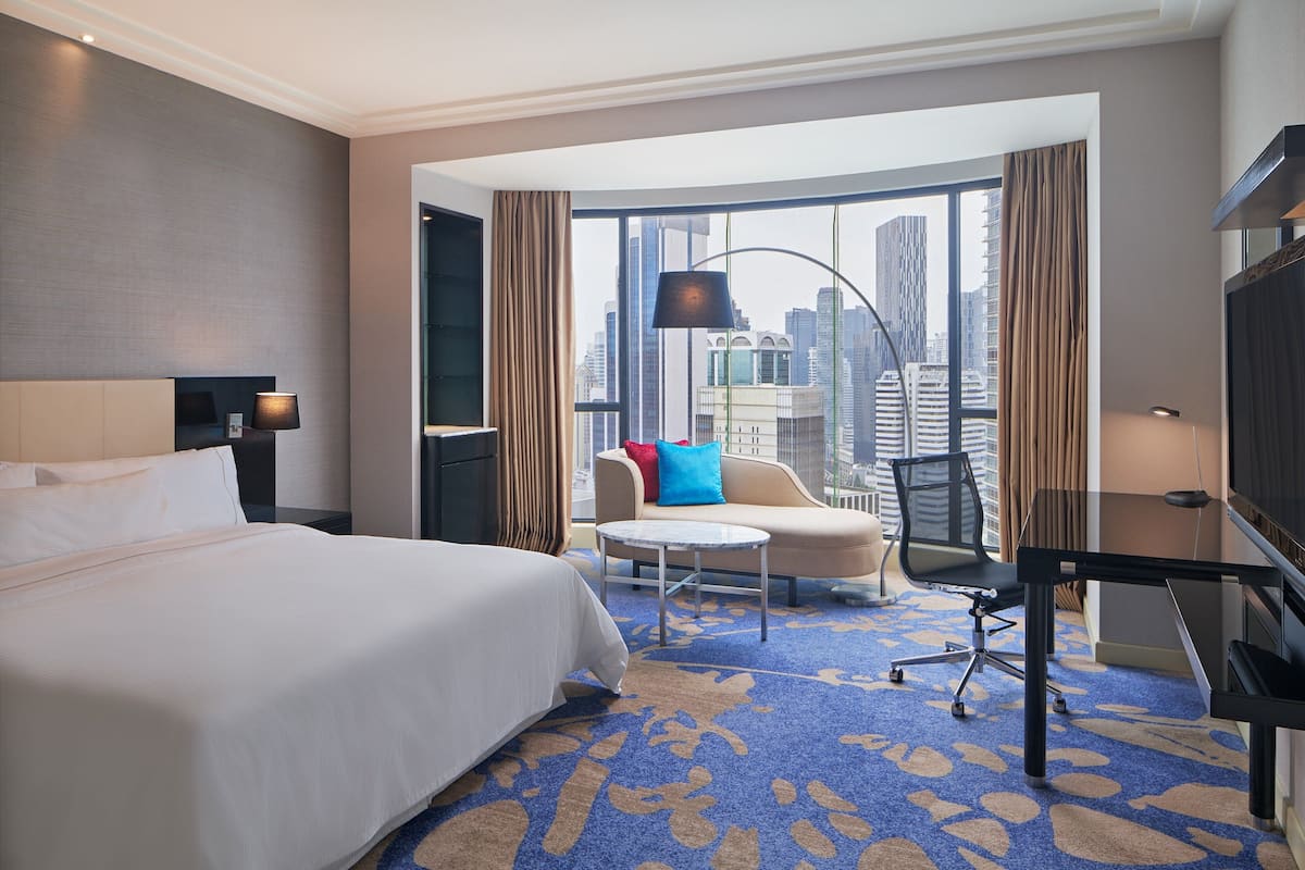 A hotel room with a large bed, a desk, a chair, a small table, and a view of the cityscape through large windows.
