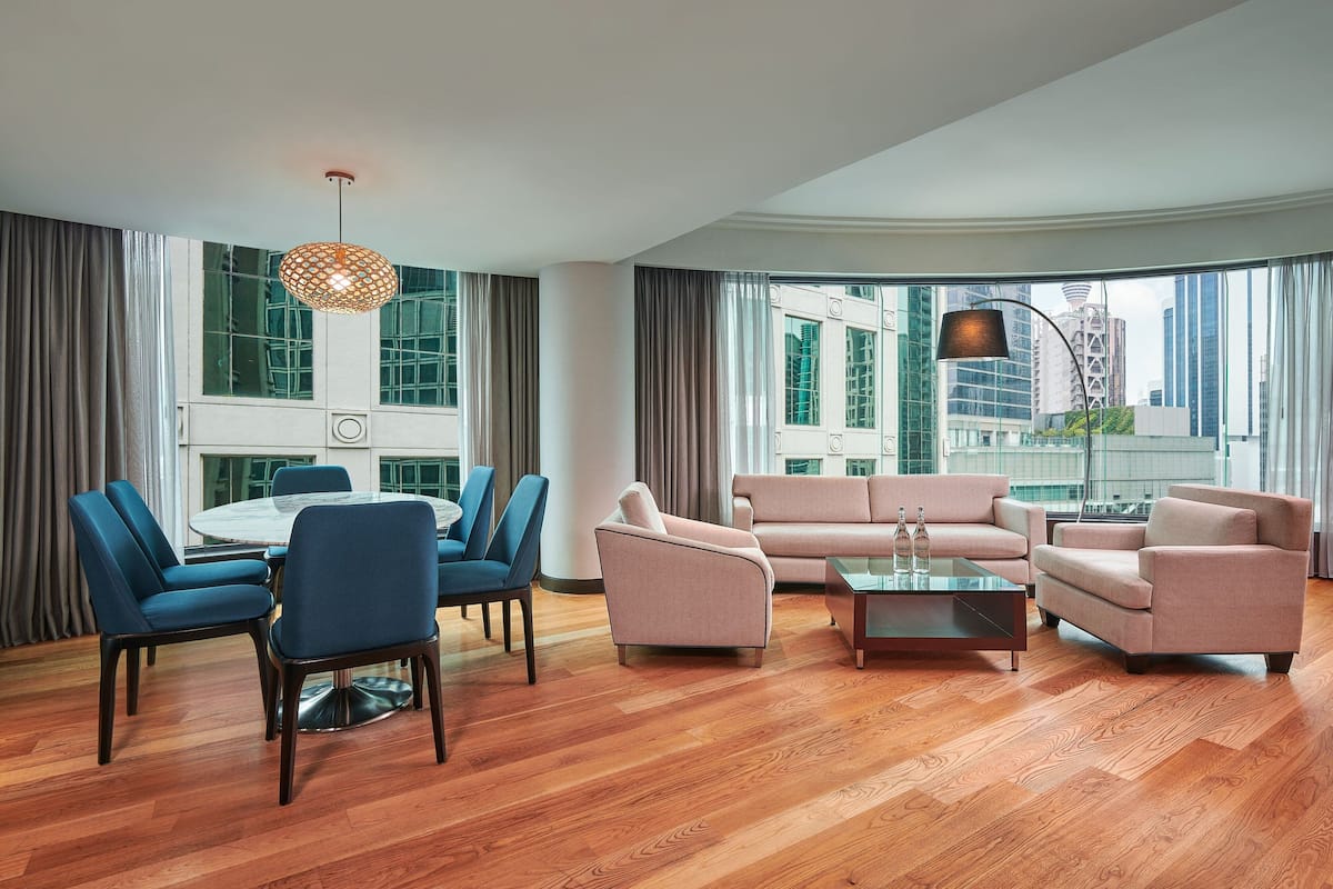 A modern living room with a round table, blue and pink chairs, a sofa set, and large windows offering a city view.