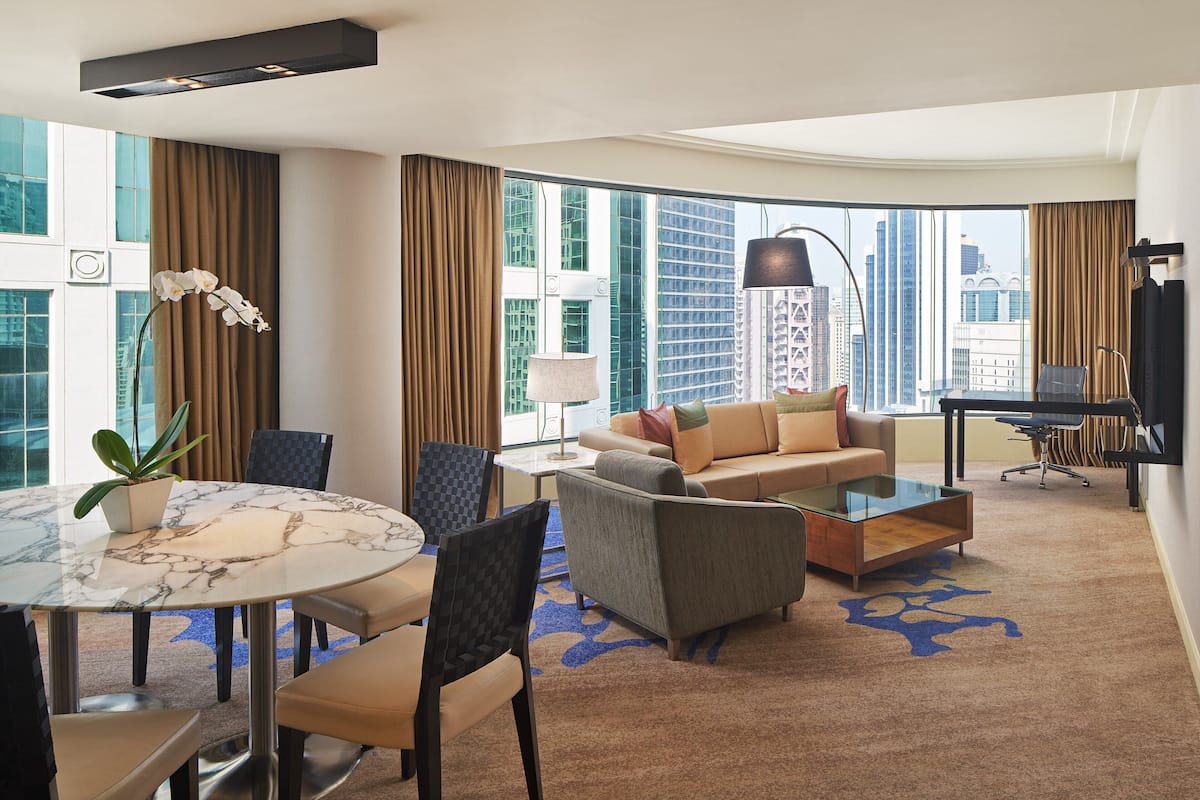 A modern hotel room with a marble dining table, a sofa, a coffee table, and a large window offering a city view.