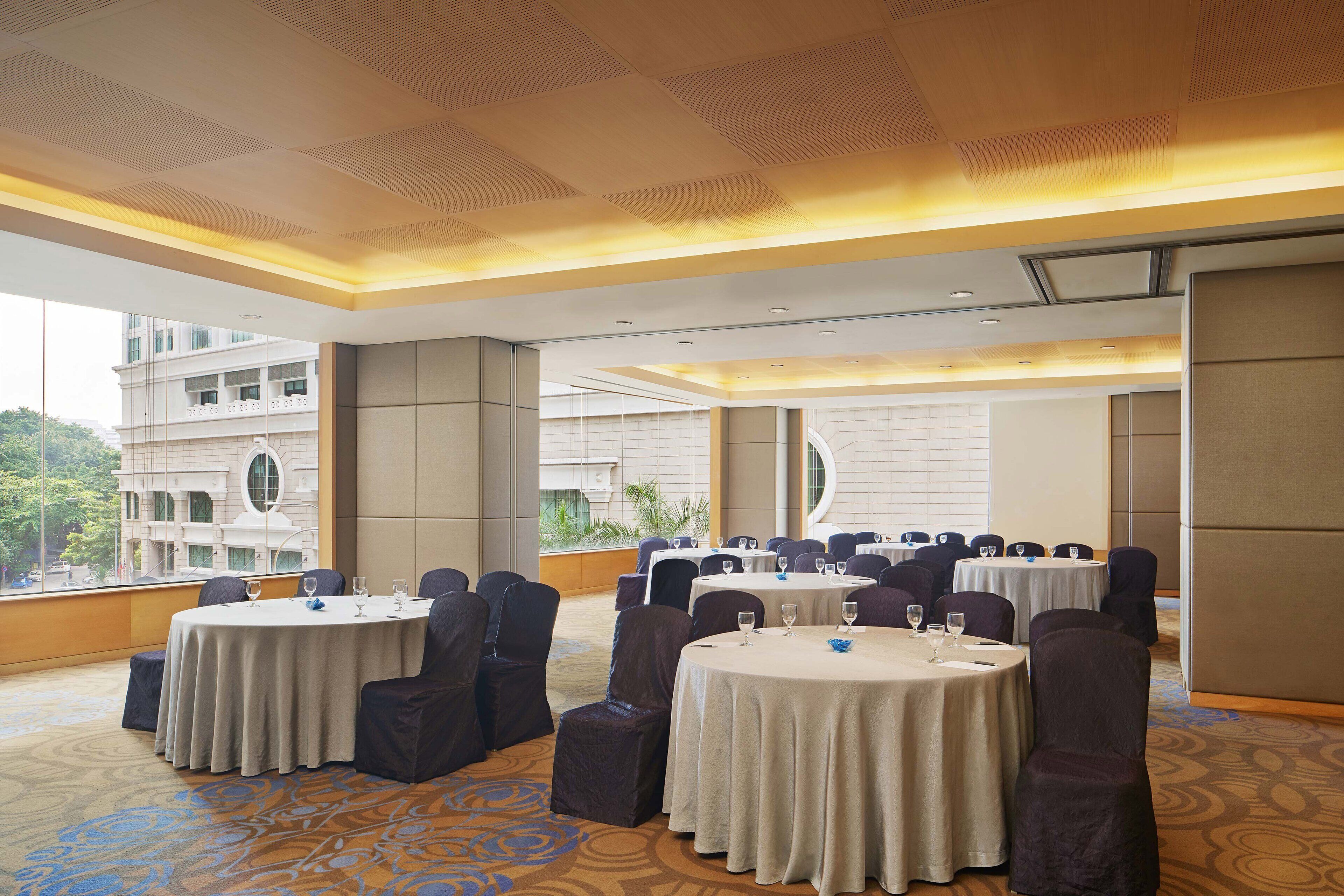 A conference room with round tables, dark chairs, and a large window offering a view of a building and greenery.