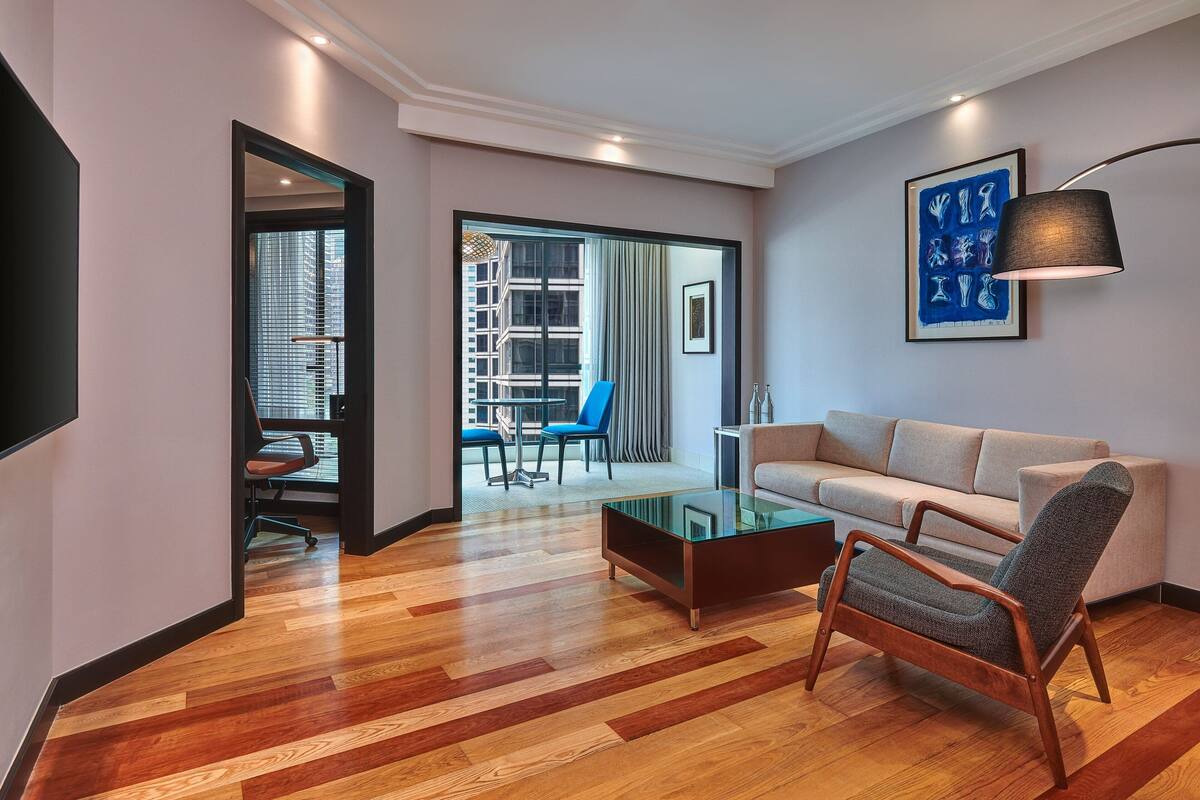 A modern living room with a glass coffee table, a sofa, and a chair. There is a framed artwork on the wall and a lamp on the side.