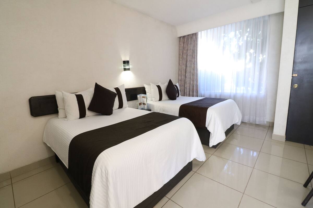 A hotel room with two beds, a nightstand, a wall-mounted lamp, and a large window with curtains.