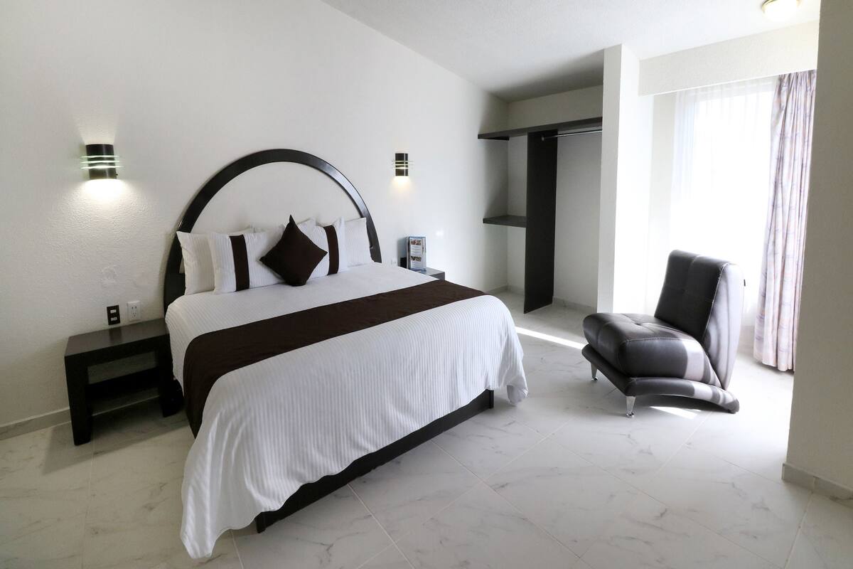 A modern hotel room with a large bed, a chair, a nightstand, and a wardrobe.