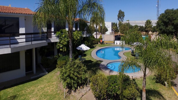 A pool area with a curved design, surrounded by palm trees and a building with a tiled roof. - Misión Express Aguascalientes Zona Norte (Jesús María)