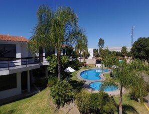 A pool area with a curved design, surrounded by palm trees and a building with a tiled roof. - Misión Express Aguascalientes Zona Norte (Jesús María)