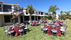 An outdoor dining area with multiple tables set for guests, surrounded by a well-maintained lawn and a building with a tiled roof. - Misión Express Aguascalientes Zona Norte (Jesús María)